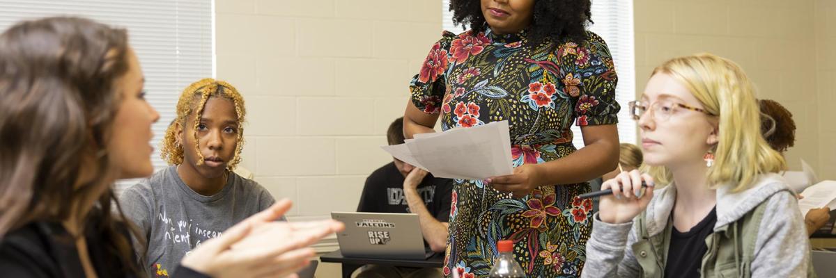 Dr. Johnson standing in her class amongst her students who are all sitting at their desks in a circle while one student has an active discussion with her. 